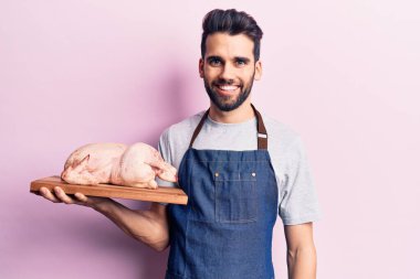 Sakallı, önlük ve çiğ tavuk taşıyan yakışıklı genç adam pozitif ve mutlu görünüyor. Kendine güvenen bir gülümsemeyle dişlerini gösteriyor. 