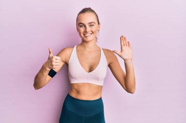 Güzel sarışın kadın spor giyiniyor ve kol bandı takıyor. Altıncı parmaklarını gösterip işaret ederken kendinden emin ve mutlu bir şekilde gülümsüyor.. 
