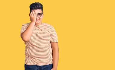 Günlük kıyafetler ve gözlük takan küçük bir çocuk bir gözünü eliyle kapatıyor, yüzünde kendine güvenen bir gülümseme ve sürpriz bir duygu var.. 