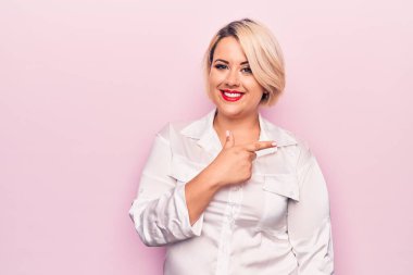 Genç, güzel, sarışın, uzun boylu, zarif bir gömlek giyen, pembe arka planda, neşeli, eli ve parmağı yana doğru gösteren bir kadın.