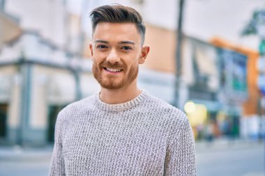 Genç beyaz adam gülümseyerek şehirde mutlu mesut duruyor..