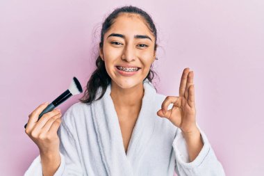 Diş teli takan, elinde makyaj fırçası olan İspanyol bir genç kız parmaklarıyla imza atıyor, gülümsüyor ve mükemmel bir sembol oluşturuyor. 