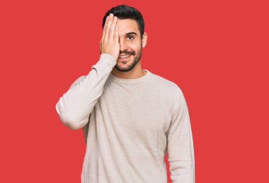 Latin asıllı genç adam gündelik giysiler giyiyor. Bir gözünü eliyle kapatıyor. Yüzünde kendine güvenen bir gülümseme ve sürpriz bir duygu var.. 