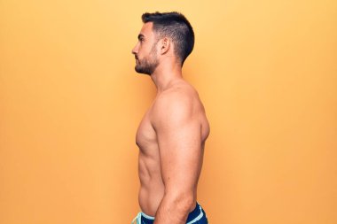 Sakallı, kolsuz tişörtlü genç adam sarı arka planda duruyor, yan yana bakıyor, rahat poz veriyor, kendine güvenen bir gülümsemesi var..