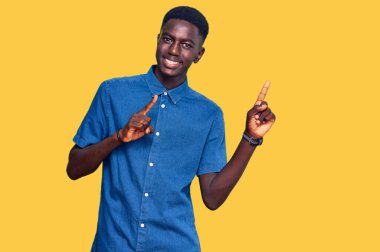 Genç Afro-Amerikalı sıradan kıyafetler giyen, gülümseyen ve kameraya bakan iki elini ve parmağını yana doğru gösteren bir adam.. 