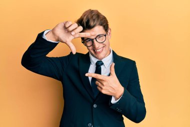 İş elbisesi ve kravat takan yakışıklı beyaz adam gülümseyen eller ve mutlu yüzlü parmaklarla çerçeve yapıyor. yaratıcılık ve fotoğrafçılık kavramı. 