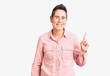 Young woman with short hair wearing casual clothes with a big smile on face, pointing with hand finger to the side looking at the camera. 
