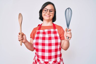 Esmer kadın, aşçı fırçası ve kaşıkla yemek pişirirken yüzünde mutlu ve havalı bir gülümsemeyle gülümsüyor. diş gösterme. 