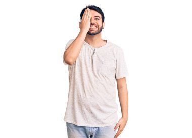 Latin asıllı genç adam gündelik giysiler giyiyor. Bir gözünü eliyle kapatıyor. Yüzünde kendine güvenen bir gülümseme ve sürpriz bir duygu var.. 