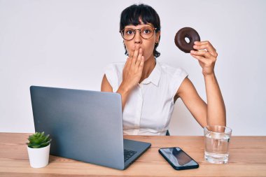 Ofiste çalışan güzel esmer kadın çikolatalı donut yerken ağzını eliyle kapadı, şok oldu ve hata yapmaktan korktu. şaşırmış ifade 