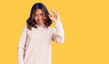 Balıkçı yaka kazak giyen genç, güzel, melez bir kadın gülümsüyor ve el ve parmaklarıyla 