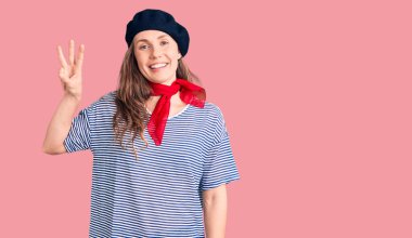 Fransız beresi ve çizgili tişört giyen genç ve güzel sarışın kadın üç numaralı parmaklarıyla kendini beğenmiş ve mutlu gülümserken onu gösteriyor.. 