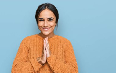 Genç esmer kadın, sıradan bir kazak giyip ellerinin birarada dua ederek af diliyor kendinden emin bir şekilde gülümsüyor.. 