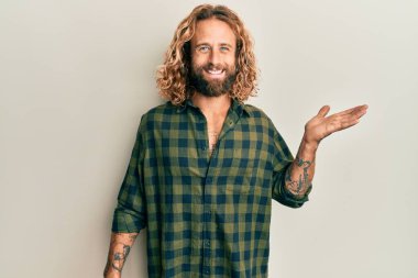 Sakallı, uzun saçlı, sıradan kıyafetler giyen, neşeli bir şekilde gülümseyen ve avuç içi kameraya bakan yakışıklı bir adam.. 