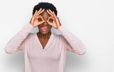 Genç Afro-Amerikan kız günlük kıyafetler giyiyor dürbün gibi jest yapıyor, dili dışarı çıkarıyor, gözleri parmaklarından bakıyor. çılgın ifade. 