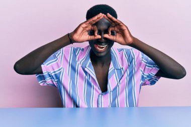 Genç Afro-Amerikan bir adam, günlük giysiler içinde masada oturmuş dürbün gibi jest yapıyor, gözleri parmaklarından bakıyor. çılgın ifade. 