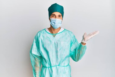 Cerrah üniforması ve tıbbi maske takan genç yakışıklı adam mutlu bir şekilde gülümsüyor ve avuç içi kameraya bakıyor.. 
