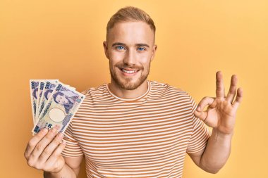 Elinde Japon Yen banknotları tutan genç beyaz adam parmaklarıyla imza atıyor, gülümseyerek mükemmel bir sembol oluşturuyor. 