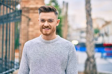 Genç beyaz adam gülümseyerek şehirde mutlu mesut duruyor..