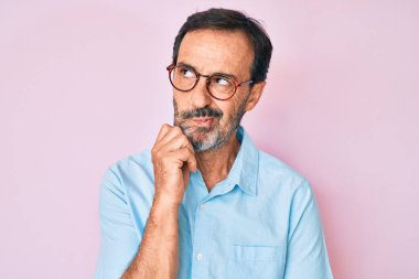 Orta yaşlı İspanyol bir adam günlük giysiler ve gözlük takıyor şüpheye odaklanmış, çenesine parmak geçirmiş ve kafasını kaldırıp merak ediyor. 