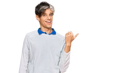 Gündelik kıyafetler giyen, gülümseyen, gülümseyen ve başparmağıyla yan tarafı gösteren İspanyol genç adam.. 