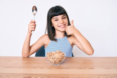 Bangır bangır oturmuş mısır gevreği yiyor mutlu ve pozitif gülümsüyor, başparmağını kaldırıyor ve onay işareti yapıyor. 