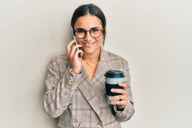 Genç esmer kadın akıllı telefon kullanıyor ve bir fincan kahve içiyor mutlu ve havalı bir gülümsemeyle gülümsüyor. diş gösterme. 