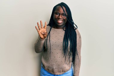 Saçları örgülü, günlük kıyafetler giyen, gözlüklü, dört numaralı parmaklarıyla işaret eden, kendinden emin ve mutlu bir şekilde gülümseyen genç zenci kadın.. 