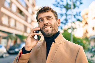 Zarif bir ceket giyen yakışıklı bir iş adamı akıllı telefon kullanarak dışarıda mutlu bir şekilde gülümsüyor.