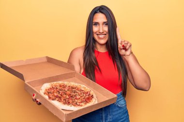 Young beautiful brunette woman holding delivery box with italian pizza smiling with an idea or question pointing finger with happy face, number one 