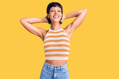 Kısa saçlı genç esmer kadın rahat ve esnek giyiniyor, kolları ve elleri başının arkasında mutlu bir şekilde gülümsüyor. 