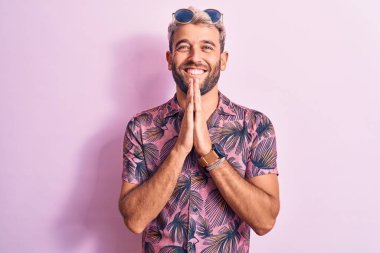 Yakışıklı sarışın, tatilde, günlük gömlek ve güneş gözlüğü takmış pembe arka planda el ele dua ediyor, af diliyor, kendinden emin bir şekilde gülümsüyor..