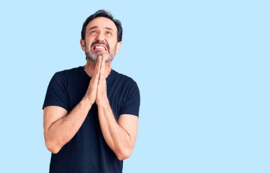 Orta yaşlı yakışıklı bir adam gündelik tişörtler giyip elleri bağlı yalvarıyor ve dua ediyor. Yüzünde umut ifadesi var. Çok duygusal ve endişeli. Yalvarıyor. 