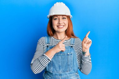Güzel kızıl saçlı kadın mimar şapkası takıyor gülümsüyor ve kameraya bakıyor iki eliyle ve parmaklarıyla kenarı işaret ediyor.. 
