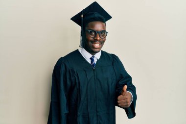 Mezuniyet şapkası ve tören cübbesi takan yakışıklı siyahi adam mutlu bir el hareketi yapıyor. Kameraya bakıp başarıyı gösteren ifade. 