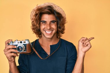 Şapka takan İspanyol genç adam, elinde klasik kamera, gülümsüyor, mutlu bir şekilde elini ve parmağını yana doğru gösteriyor. 