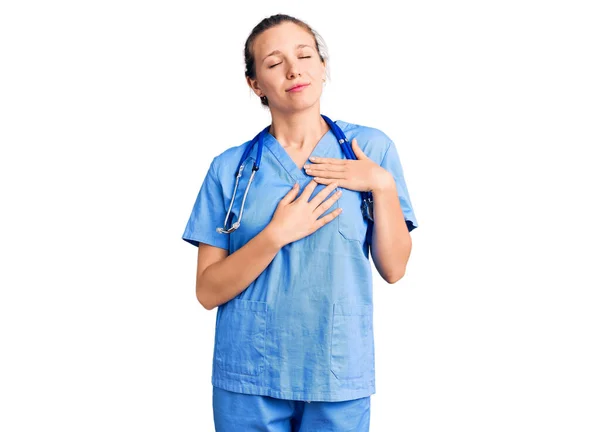 医師の制服を着た若い美しいブロンドの女性と目を閉じて胸に手で笑みを浮かべて聴診器と顔に感謝のジェスチャー 健康の概念 — ストック写真