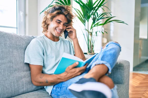 Young hispanic man smiling happy talking on the smartphone and reading book at home