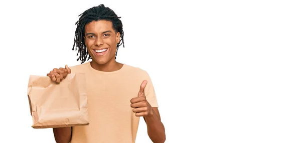 若いアフリカ系アメリカ人の男を持って紙の袋を奪う笑顔幸せと肯定的な 親指アップ行う優れた承認サイン — ストック写真