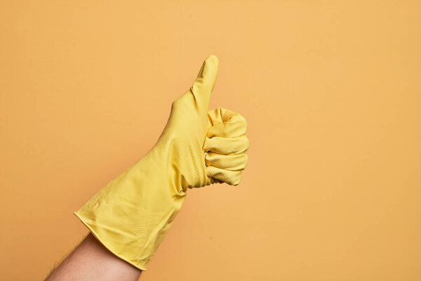 Hand of caucasian young man with cleaning glove over isolated yellow background doing successful approval gesture with thumbs up, validation and positive symbol
