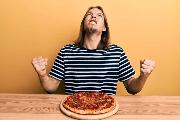 英俊的白种人 留着长发 疯狂地吃着美味的意大利香肠披萨 疯狂地喊叫着 带着咄咄逼人的表情和手臂大喊大叫 沮丧的概念 — 图库照片
