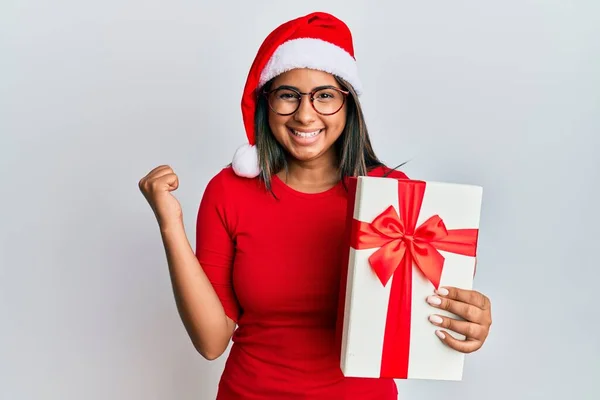 Young Latin Girl Wearing Christmas Hat Holding Gift Screaming Proud — 스톡 사진