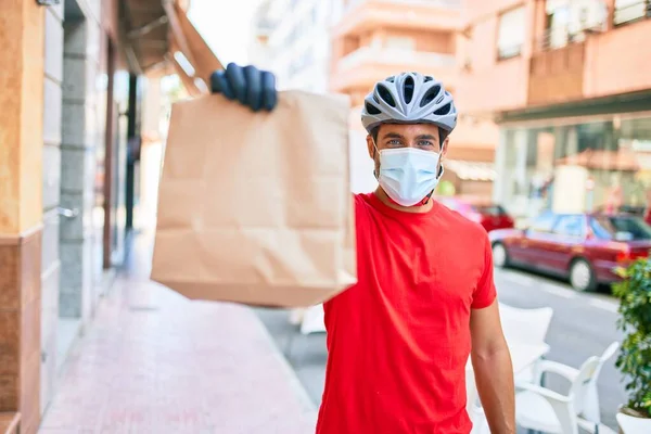 若い配達人は自転車のヘルメットとコロナウイルス保護医療用マスクを着て街の通りで紙袋を奪う — ストック写真