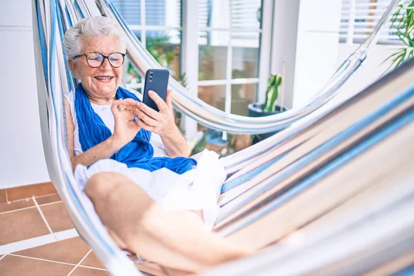 Elder senior woman with grey hair smiling happy relaxing on a hammock at home using smartphone