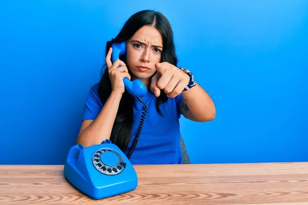 Beautiful brunette young woman speaking on vintage telephone pointing with finger to the camera and to you, confident gesture looking serious