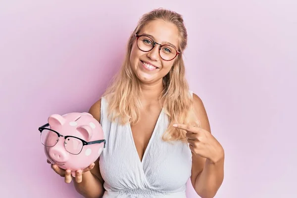 Jong Blond Meisje Holding Piggy Bank Met Bril Wijzend Vinger — Stockfoto