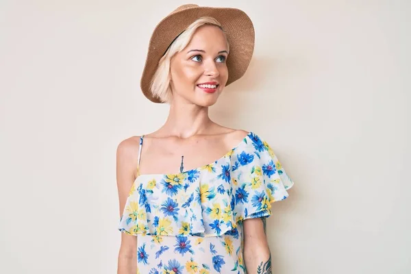 Young blonde woman with tattoo wearing summer hat looking away to side with smile on face, natural expression. laughing confident.