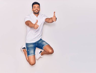 Genç, yakışıklı, sıradan kıyafetler giyen, mutlu gülümseyen bir İspanyol. Yüzünde tebessümle zıpla, tamam işareti yap, başparmağınla izole beyaz arkaplan üzerinde.