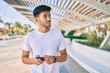 Genç Latin adam mutlu bir şekilde gülümsüyor ve şehirde akıllı telefon kullanıyor..