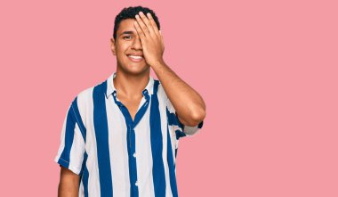 Gündelik giysiler giyen genç bir Arap bir gözünü eliyle kapatıyor, yüzünde kendine güvenen bir gülümseme ve sürpriz bir duygu var.. 